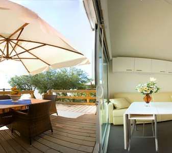 Area con terrazza in legno, ombrellone grande e tavolo, accanto a salotto moderno con divano beige.