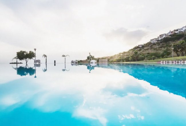 Una piscina a sfioro riflette il cielo, circondata da colline con edifici bianchi.