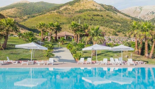 Piscina con lettini e ombrelloni bianchi, circondata da palme e colline verdi sullo sfondo.