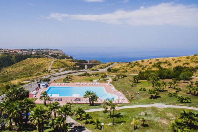 Piscina all'aperto con lettini, circondata da colline verdi e vista sul mare.