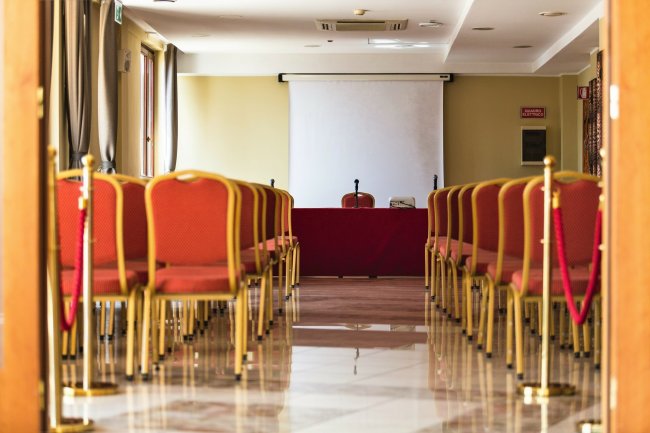 Sala conferenze vuota con sedie rosse e schermo bianco.