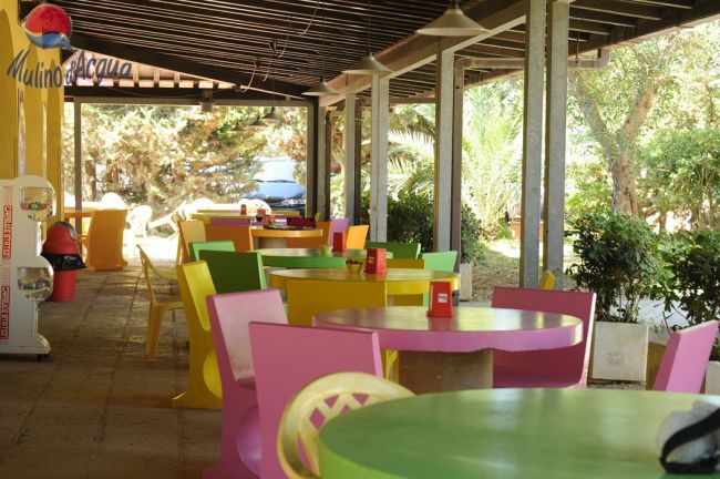 Terrazza esterna di un caffè con sedie e tavoli colorati, circondata da alberi.