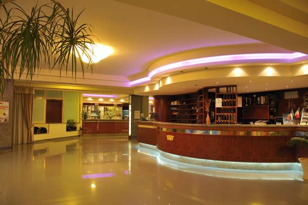 Reception di un hotel con banco in legno curvo, luci soffuse colorate e piante decorative.