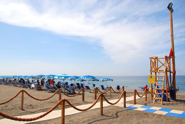 Spiaggia sabbiosa con ombrelloni blu e bagnanti, vista torretta di legno.