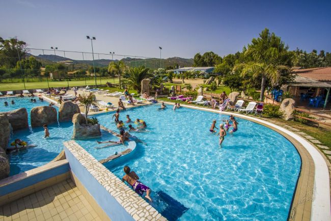 Piscina all'aperto affollata con giochi d'acqua, circondata da lettini e vegetazione.
