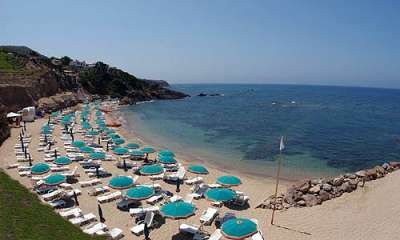 Spiaggia con sabbia chiara, ombrelloni turchesi e sdraio. Mare calmo e rocce sullo sfondo.