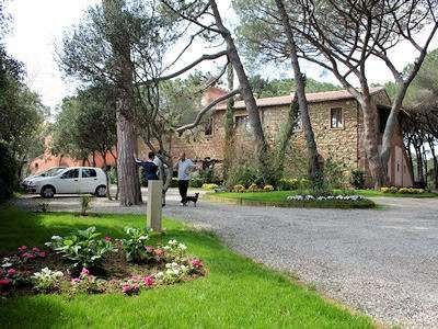 Strada sterrata con area giardino fiorita, due persone e un edificio in pietra tra gli alberi.
