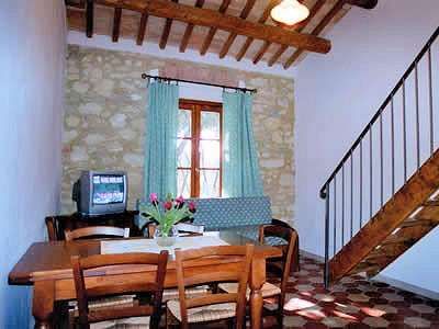 Sala con parete in pietra, tavolo, sedie, divano, finestra con tende, e scala in legno.