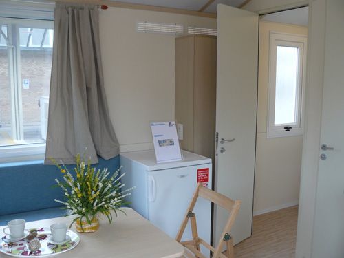 Un interno luminoso con tavolo apparecchiato, una finestra, frigo e porta aperta.