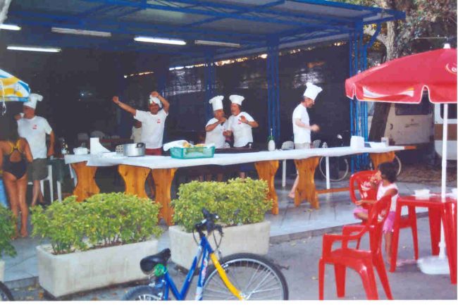 Cucinieri in divisa preparano cibo sotto una tettoia, decorata con sedie rosse e una bici.