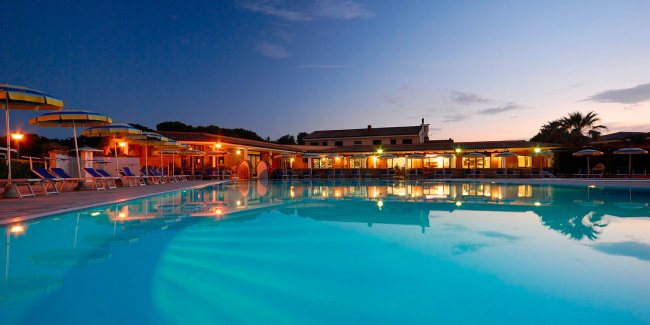 Piscina illuminata di sera con ombrelloni e sedie a sdraio.