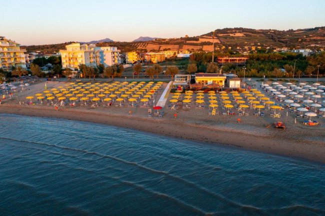 Vista aerea di una spiaggia con ombrelloni gialli, edifici e colline sullo sfondo.