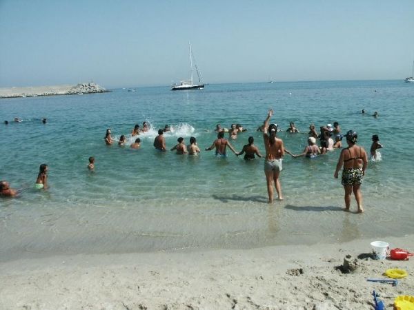 Spiaggia con persone che giocano in acqua, barca a vela in lontananza.