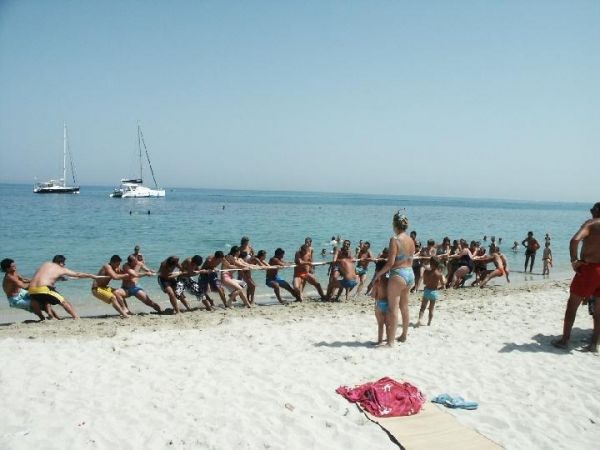 Persone giocano al tiro alla fune su una spiaggia di fronte al mare con barche a vela in lontananza.
