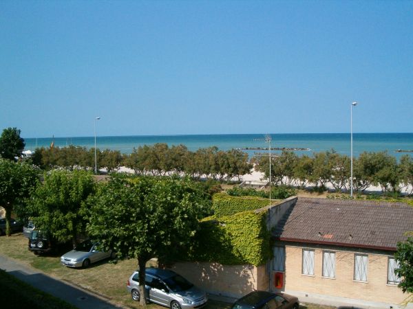 Vista sul mare con una casa in primo piano, alberi e auto parcheggiate lungo la strada.