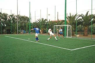 Partita di calcio su campo verde recintato, con due giocatori vicino alla porta e un portiere.