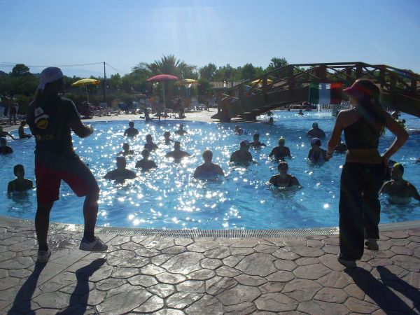 Persone partecipano a una lezione di acquagym in piscina con due istruttori in primo piano.