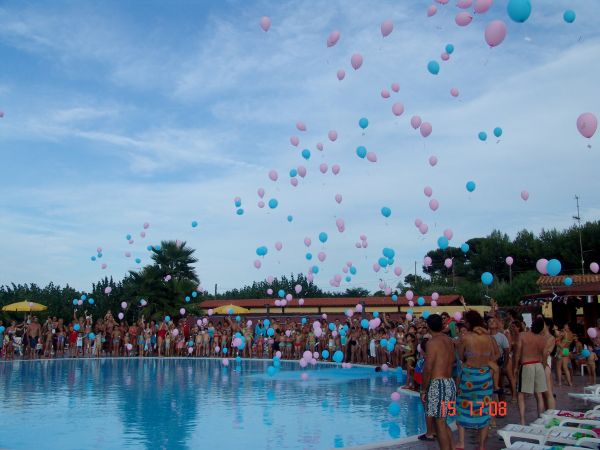 Persone intorno a una piscina guardano palloncini rosa e blu che volano in cielo.