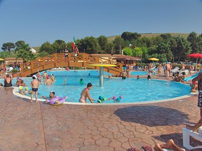 Piscina all'aperto con molte persone che nuotano e giocano, un ponte di legno al centro.