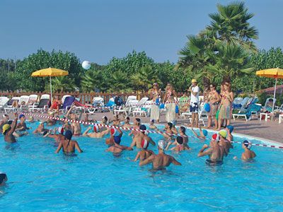 Persone giocano a pallavolo in piscina con ombrelloni gialli e palme sullo sfondo.