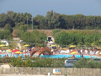 Vista di una piscina all'aperto con ombrelloni gialli e un ponte in legno, alberi e siepi intorno.