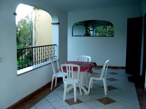 Terrazza con tavolo rotondo coperto da tovaglia rossa e quattro sedie bianche, vista su alberi.