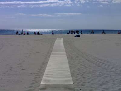 Spiaggia sabbiosa con passerella di legno che conduce al mare, persone in lontananza.