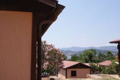 Vista parziale di edifici beige con tetto rosso, alberi e montagne sullo sfondo.
