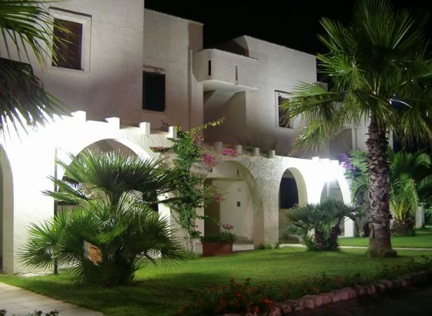 Edificio bianco illuminato di notte, circondato da palme e giardino verde.