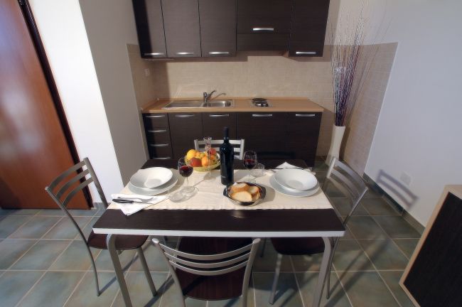 Tavolo apparecchiato con piatti, vino e frutta, cucina sullo sfondo.
