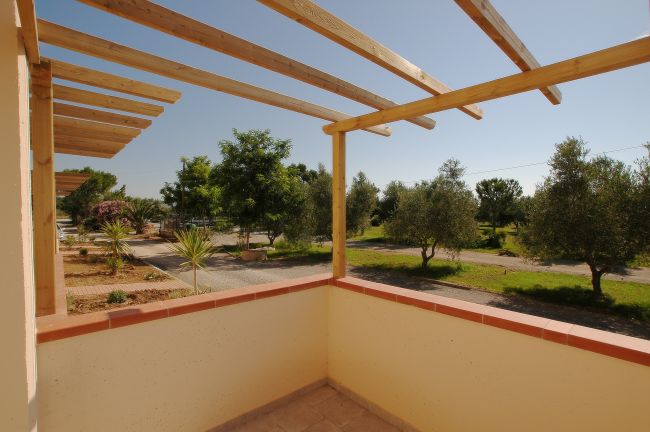 Balcone con pergolato in legno, vista su un giardino con alberi e cielo sereno.