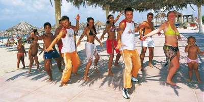 Un gruppo di persone balla sulla spiaggia sotto ombrelloni di paglia.