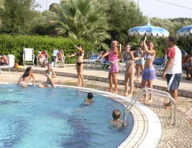 Persone intorno a una piscina, alcune in piedi, altre in acqua.