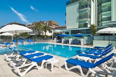 Piscina all'aperto con lettini blu e ombrelloni bianchi, accanto a edifici moderni.