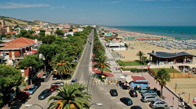 Viale alberato lungo la costa con spiaggia e ombrelloni.