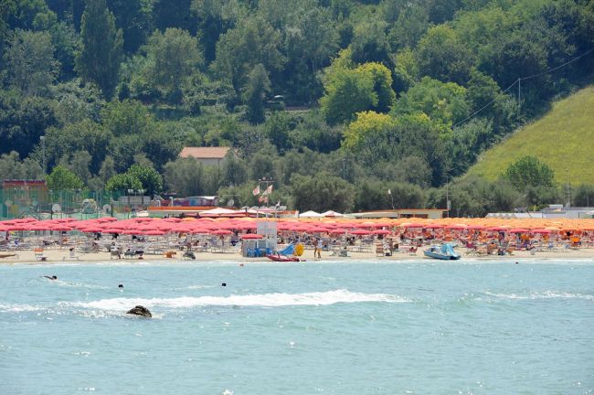 Spiaggia con numerosi ombrelloni rossi e verde vegetazione alle spalle.