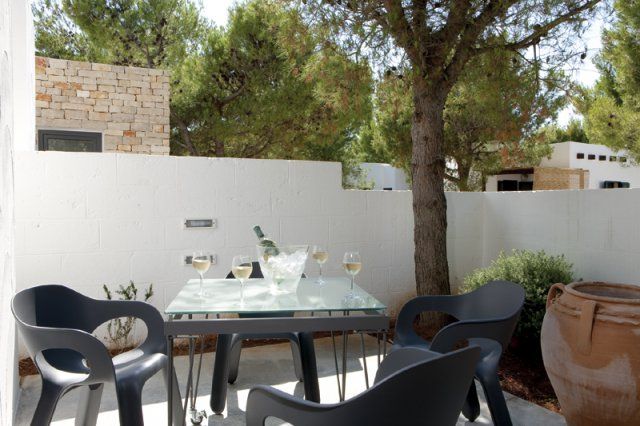 Tavolo all'aperto con quattro bicchieri di vino, circondato da alberi e un muro bianco basso.