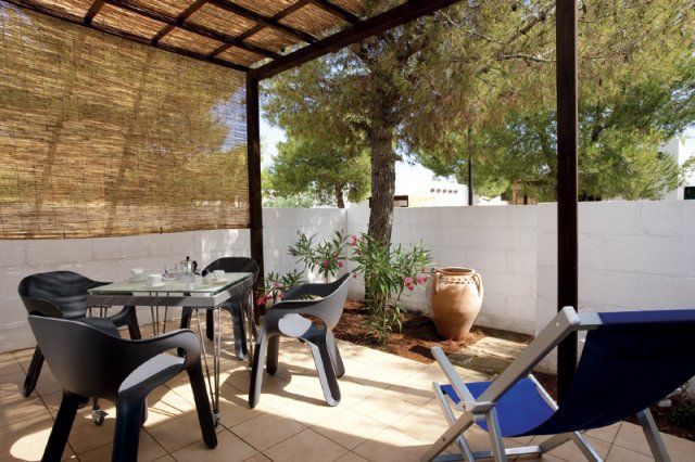 Terrazza coperta con tavolo, sedie e sdraio blu, circondata da un muro bianco e vegetazione.