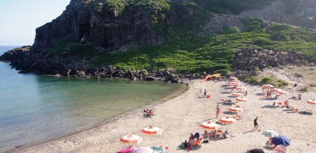 Spiaggia con ombrelloni rossi e bianchi, persone rilassate vicino ad una collina verde.