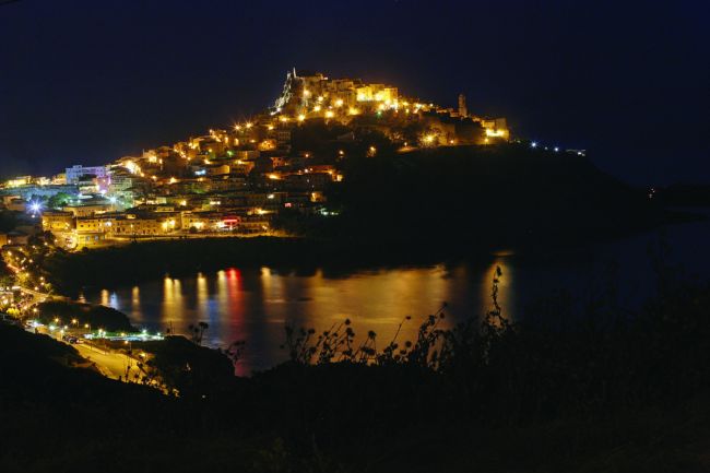 Vista notturna di una cittadella illuminata su una collina che si riflette nell'acqua.