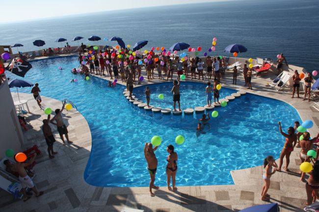 Un'affollata piscina all'aperto con persone che giocano con palloncini colorati, vista mare.