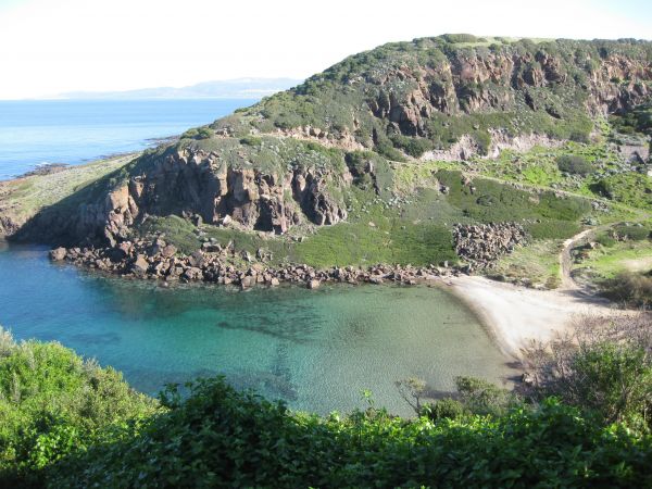 Piccola baia con spiaggia sabbiosa, circondata da colline rocciose e vegetazione.