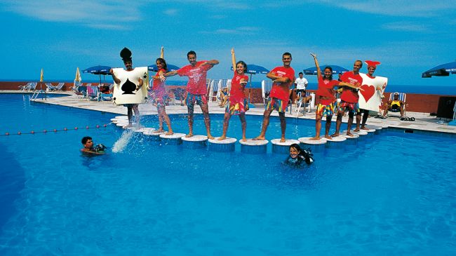 Persone in piedi su piattaforme in una piscina, alcune indossano costumi a tema carte da gioco.