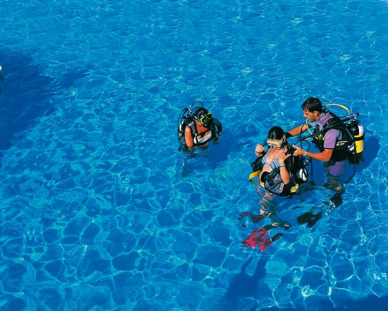 Tre persone con attrezzatura subacquea in una piscina azzurra, una persona sta assistendo le altre.