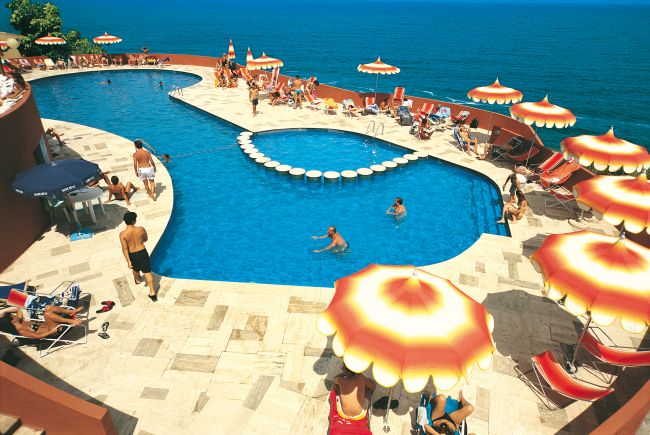 Piscina con piattaforme rotonde e ombrelloni arancioni, affacciata sul mare.