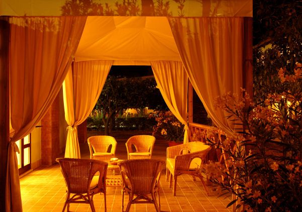 Gazebo illuminato con sedie di vimini, circondato da vegetazione.