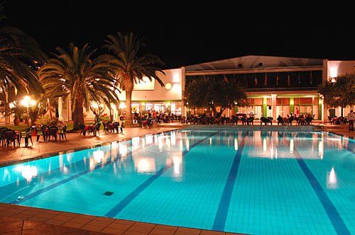 Piscina illuminata di notte con palme e un edificio sullo sfondo.