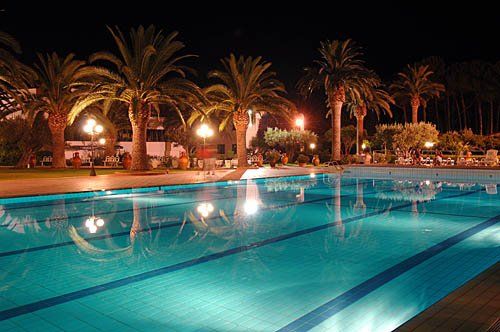 Piscina illuminata di notte con palme riflettenti in acqua.