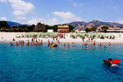 Spiaggia affollata con bagnanti e sdraio verdi, mare blu in primo piano, montagne sullo sfondo.