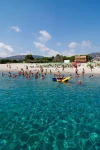 Mare limpido con persone che nuotano verso la spiaggia, ombrelloni verdi e montagne sullo sfondo.